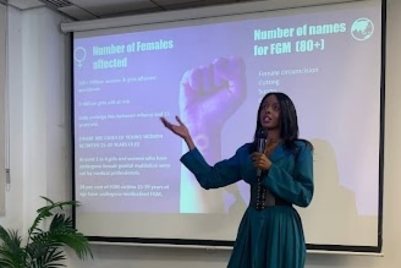 A woman in a dress stands in front of a screen with information about female genital mutilation.