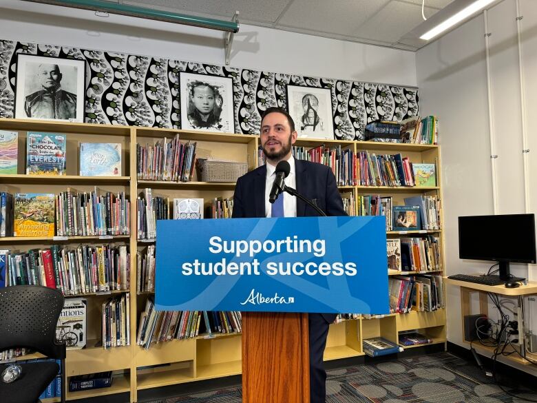 A man with dark hair and dark facial hair wearing a dark-coloured suit, white shirt, and blue tie stands behind a podium that reads 