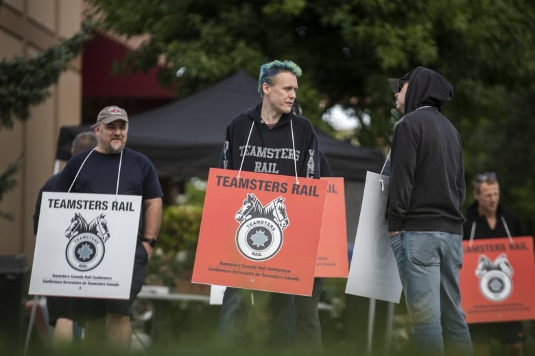 People, including a person with spikey blue-green hair, picketing and wearing signs that read: Teamsters Rail 