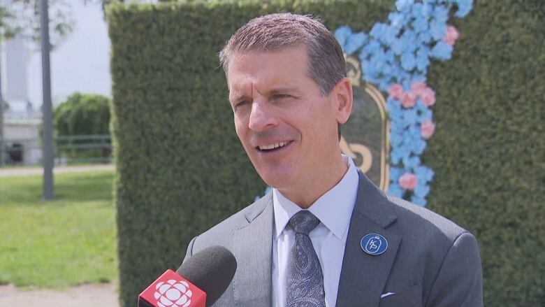 A man in a suit talks into a CBC microphone outside near a green hedge on a sunny day. He is visible from the chest up. He is white with grey hair