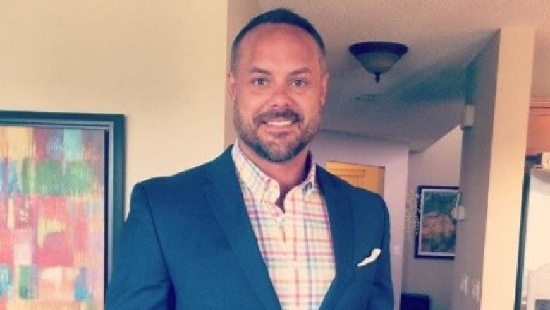 A man with short, dark and greying hair and facial hair wearing a blue suit jacket over a striped multi-coloured button up shirt.