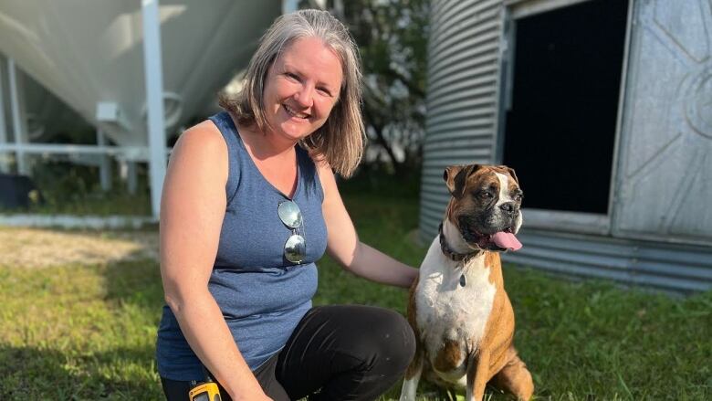 A woman kneels on the ground and smiles to the camera, with her hand on a dog sitting next to her.