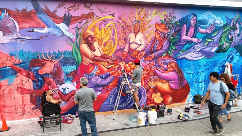 On the far left, a woman wearing a red cap sits on a black chair. She's working on a mural, but has turned around to talk with a passerby standing to her right in a grey shirt. In the middle another woman works away at the same mural on top of a ladder. Two people walk by, observing the mural on the right. 