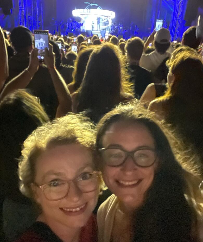 Two girls stand in concert crowd with stage behind them. 