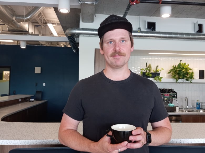 Man in black shirt and black hair holding a coffee cup. Behind him is a counter top and on a shelf if a coffee machine.