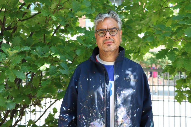 A man wearing a paint-stained navy jacket with a Chilean soccer jersey underneath poses for a picture in front of a leafy trea.