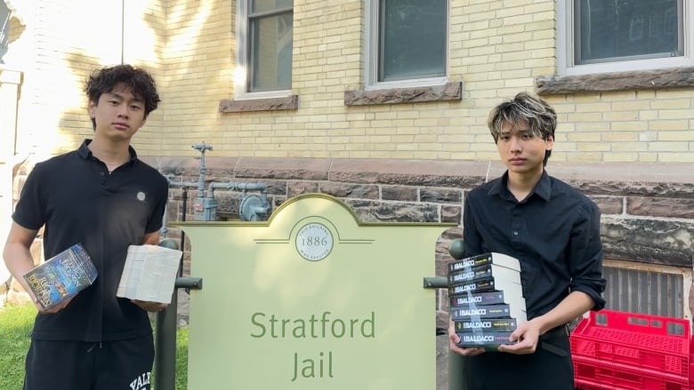 Two young men wearing black are holding stacks of books. They flank a sign reading 