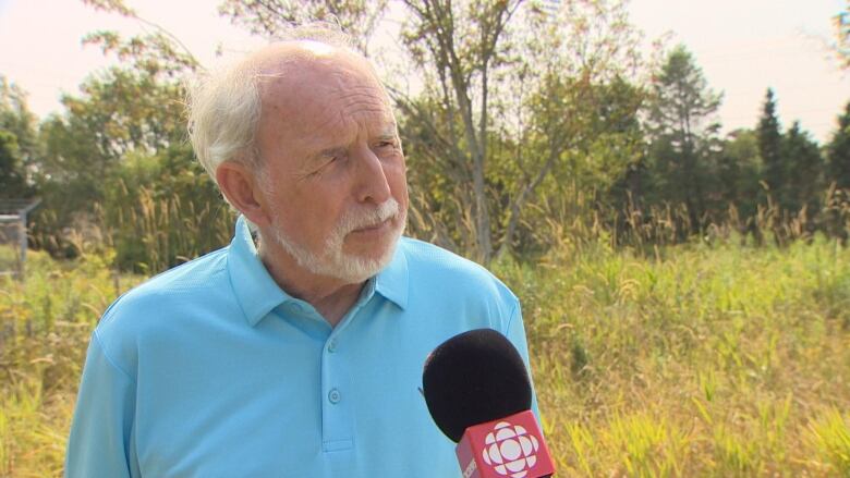 Man with white hair and blue polo shirt standing outdoors. There are trees behind him.