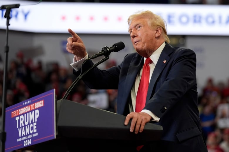 An older clean shaven man in a suit and tie points while speaking at a podium that says Trump-Vance on it. The photo is taken from a side angle, the man is seen in profile.