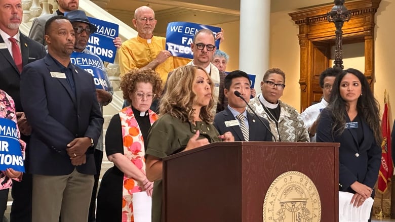 A woman is showing speaking at a podium from a distance, with a couple dozen people, both men and women, standing behind her. Some of those people hold placards whose words aren't entirely visible.