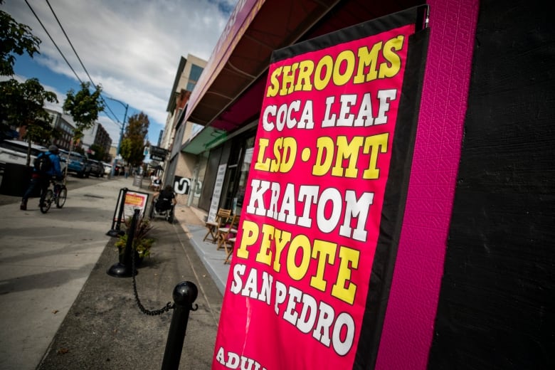 A sign reading 'Shrooms Coca Leaf LSD DMT Kratom Peyote San Pedro' is pictured on the side of a store.