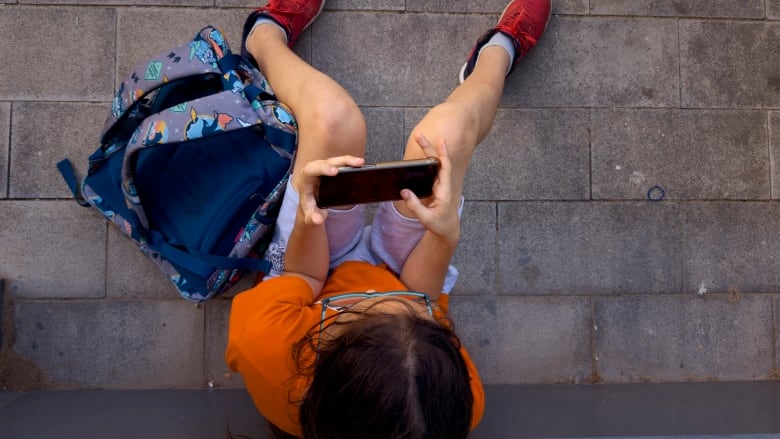 A boy with eyeglasses, wearing an orange T-shirt and red sneakers, looks at a cellphone  as he sits beside his blue knapsack.