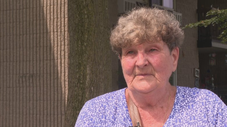 A head-shot of an elderly woman in front of the apartment building. 