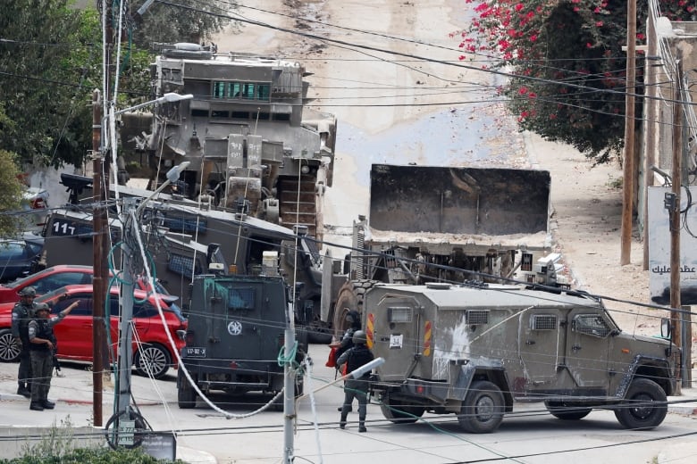 Several military vehicles are pictured on a street. A few soldiers stand nearby. 