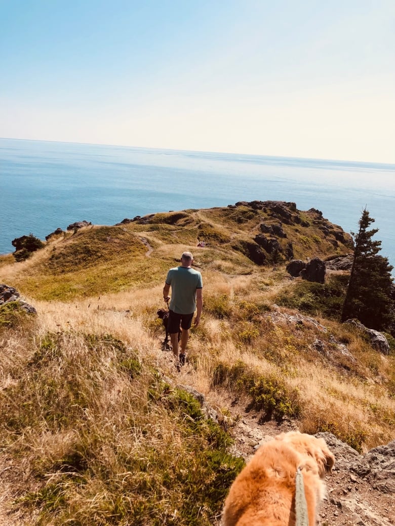 A man walks over a grassy cliff, a golden retriever dog behind him.