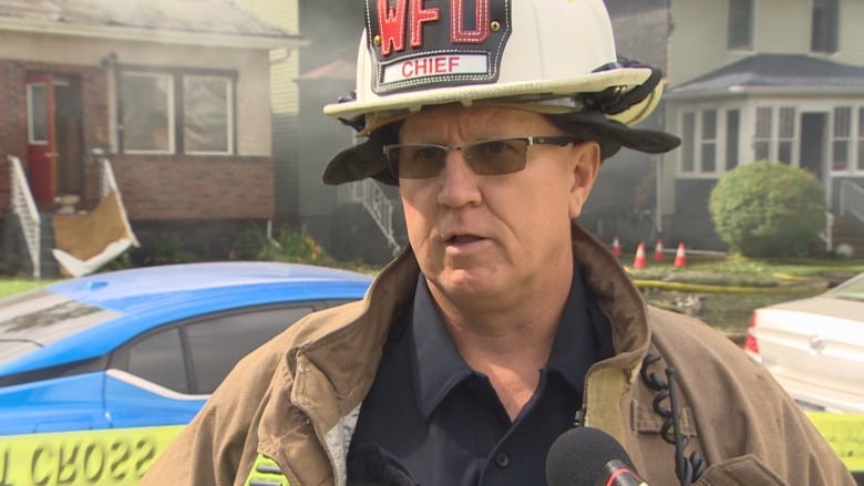 A firefighter wearing a helmet with the initials WFD stands in front of microphones on a street. Behind him are homes surrounded in smoke.