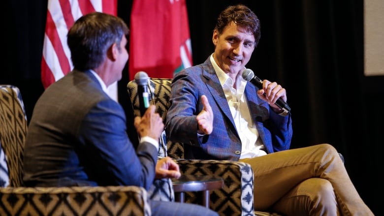 Two men converse while sitting in easy chairs on stage and holding microphones.