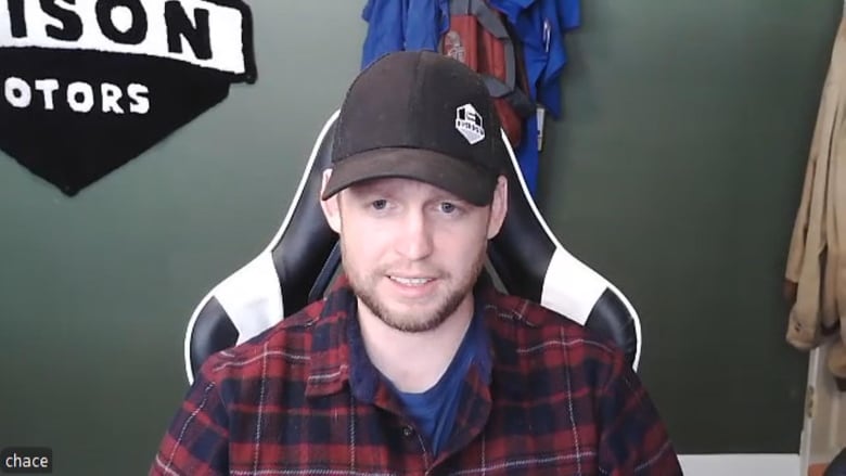 A man sitting down wearing a red and black plaid shirt and a black baseball cap. 