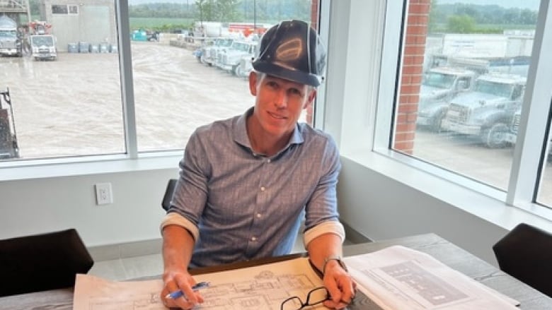 A man in a hard hat sits at a desk in a glass-walled office with a blue print.  Behind him is a gravel lot filled with parked heavy trucks.