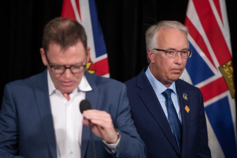 Two men speak with B.C. flags in the background. One of them is in focus and looks pensive, while the other looks down at the mic and forlorn.