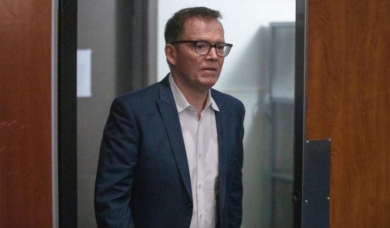 A white man looks pensive and forlorn as he enters a room through a door.