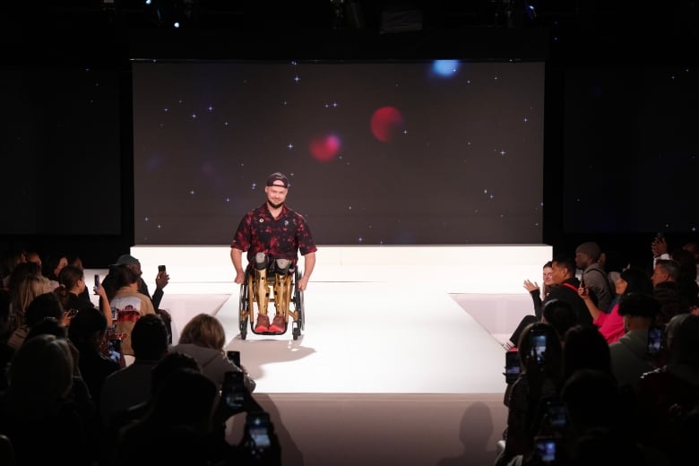 A man in a wheelchair wearing a hat is pictured on a stage with a crowd of people to the right and left of him.