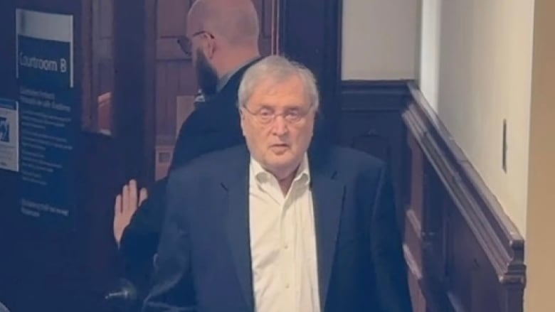 A man with white hair wearing a blazer, leaning on a cane stands in front of a metal detector at a court house in Halifax. 