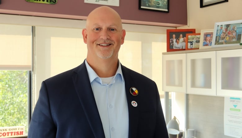 A bald man in a dark blue jacket and light blue dress shirt smiling in an office with various photos in the background.