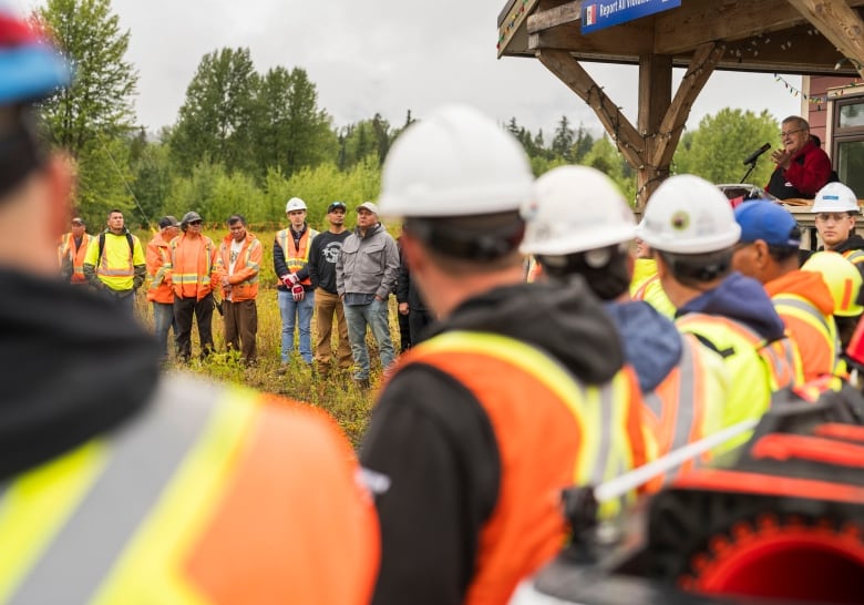 Men in hard hats and construction vests stand in a row listening to an Indigenous Elder.