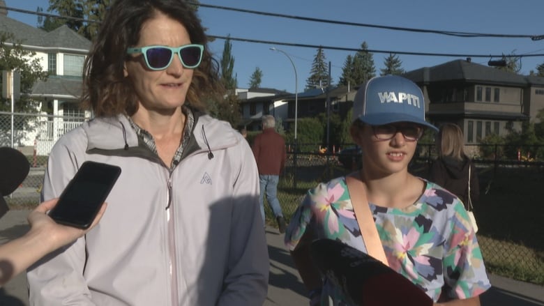 A woman with brown hair wearing sunglasses and a light-coloured windbreaker, a girl wearing a blue cap and a colourful, floral-print shirt.