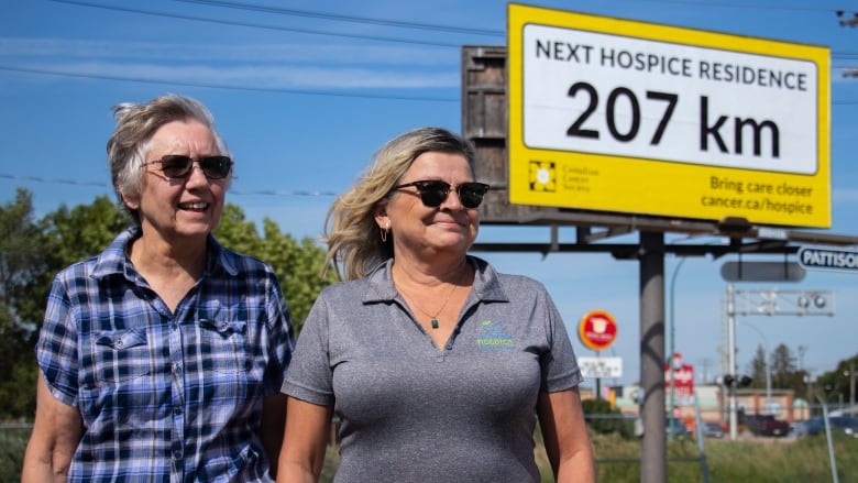 Two women stand in front of a billboard that says the nearest hospice residence is 207 kilometres away.