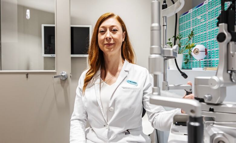 A picture of a woman with red hair, wearing a lab coat, sitting in front of equipment used to examine someone's eyes.