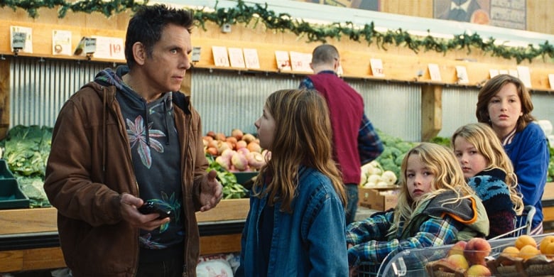 A man stands in the produce aisle of a grocery store. He appears to be speaking with a young child. Two other children sit in a grocery cart behind him, which another child is pushing. 