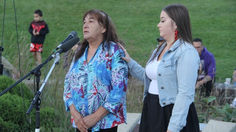 A woman in a blue blouse stands behind a microphone, beside a woman in a light blue jean jacket and white shirt underneath.