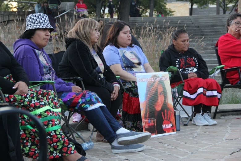 People sit in chairs, their faces solemn as their either look ahead or look down to the ground.