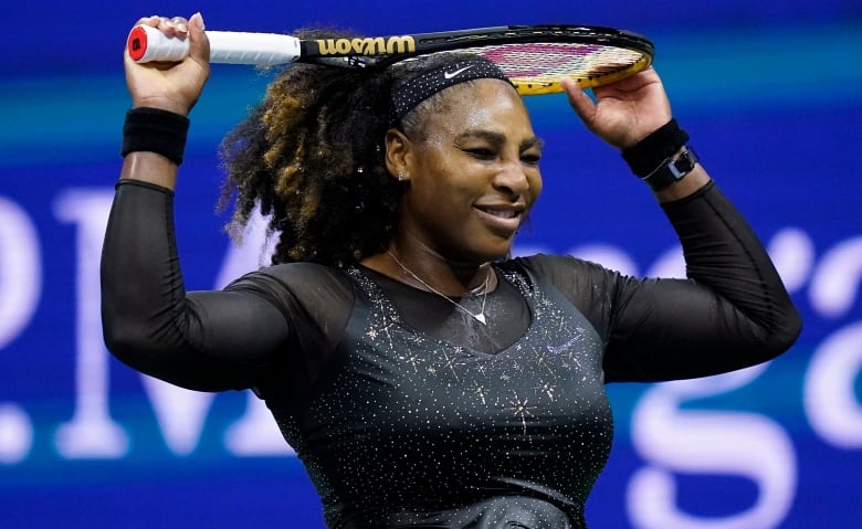 A woman smiles while resting a tennis racket on her head, one hand on the handle, the other holding the top of the racket. 