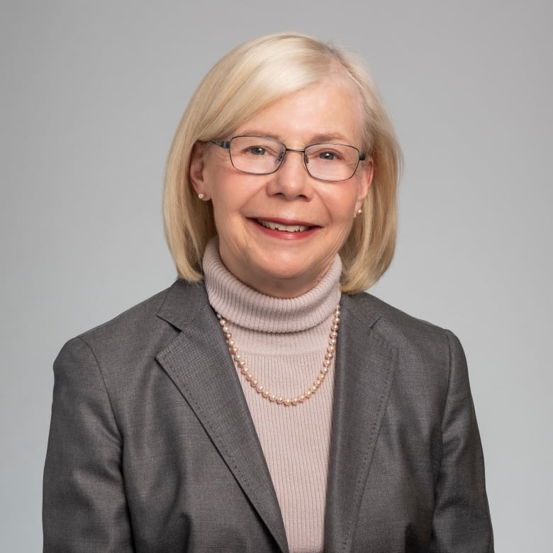 A woman with light blonde, shoulder-length hair, glasses and a grey blazer is smiling.