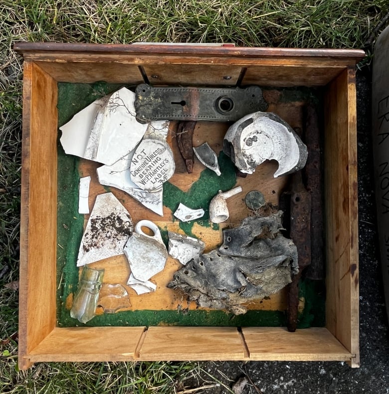 In a wooden box lie old leather from a shoe, broken pieces of ceramic, a ceramic pipe, a broken glass bottle and a keyhole cover.