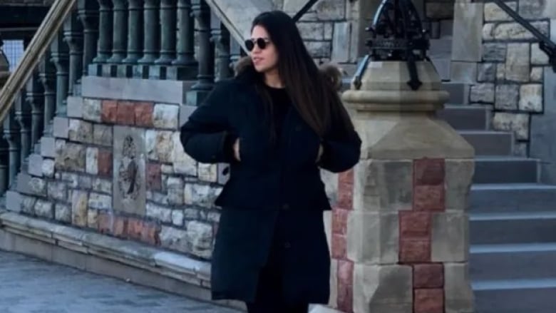 A young woman with long dark hair and sunglasses walks down a city street in front of church steps in a black winter jacket and black leggings. Her hands are in her pockets. Her head is turned toward the street. It's day time