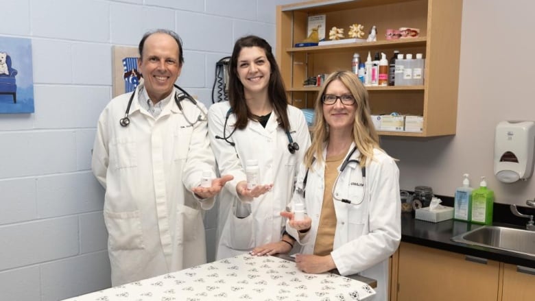 Three people in lab coats stand in front of a table holding bottles of medicine.