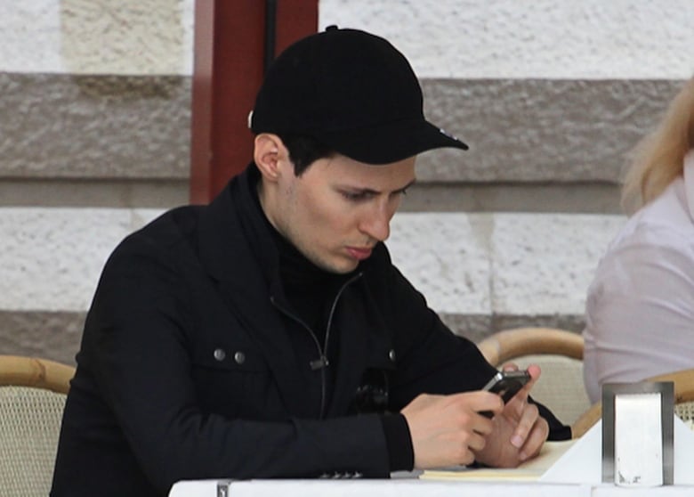 A man wearing a black ballcap, black jacket and black shirt underneath, looks at his iPhone while sitting at a table at an outdoor cafe. 