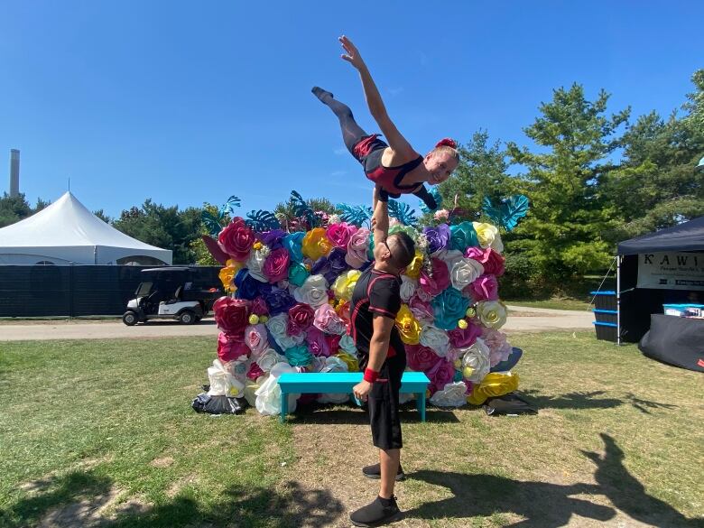 A male acrobat holding up a female acrobat with one hand in a park. 