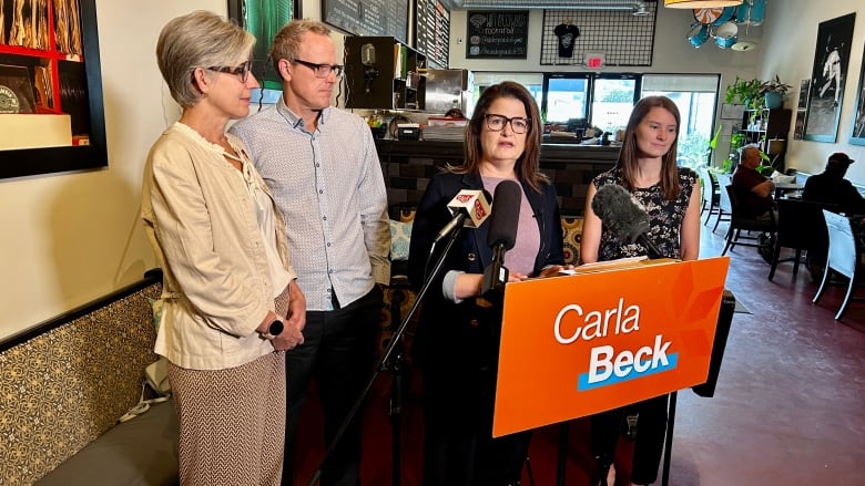 A woman wearing a purple shirt and a black jacket stands at a podium. On the podium is an orange sign with the name 