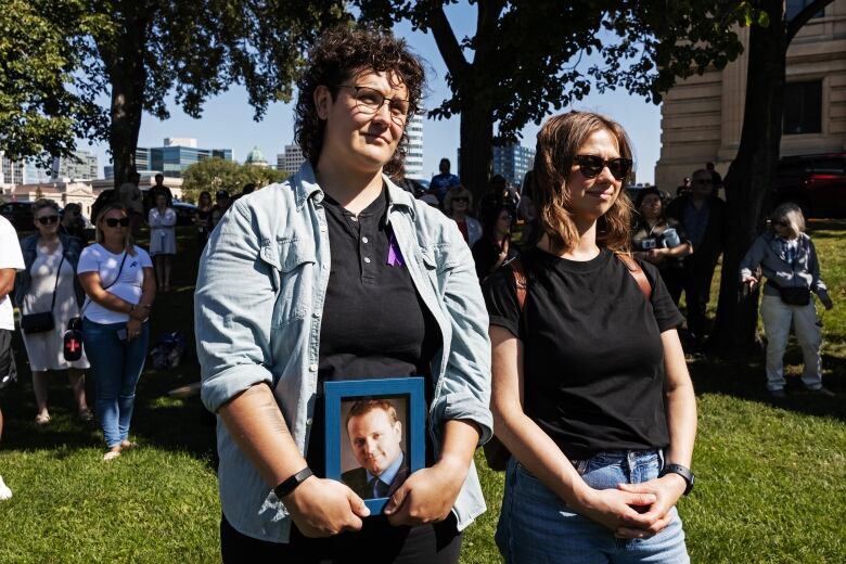 Two people stand together, with one of them holding a framed photo of a man.