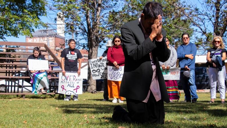 A man prays with his hands pressed together and his head down on them as he kneels in grass. People behind him hold signs to raise awareness about overdoses.