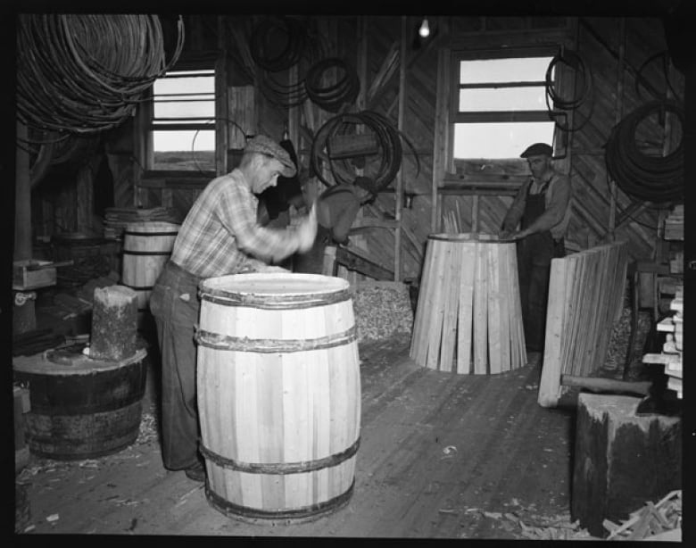 Two men work in a dark wooden workshop, making large wooden barrels. One man is caught in motion while hammering a barrel.