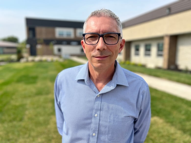 A man in a checkered blue and white shirt and blue-framed glasses stands on a lawn in front of a school.