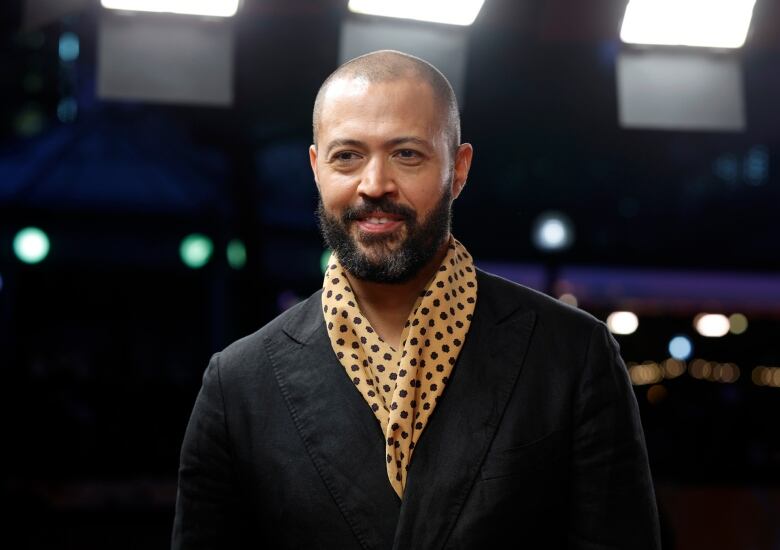 A smiling man in a scarf stands in front of a dark background. 