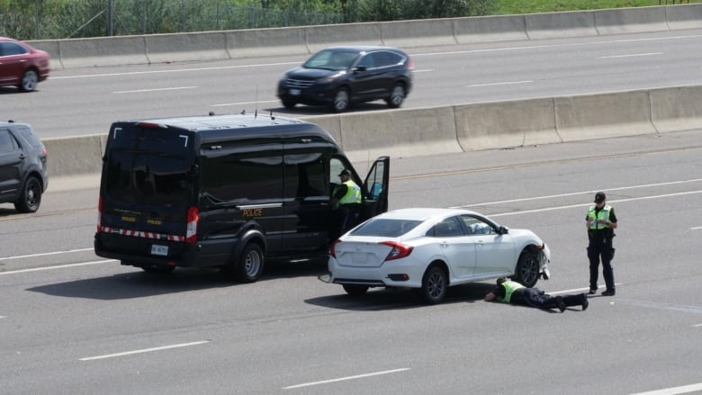 A police van is seen next to a white sedan with police officers standing next to it.