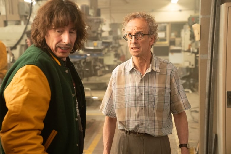 A man in a school jacket stands next to an older man in a woodworking shop a still from a film.